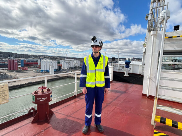 Surveyor in high‑vis vest, helmet and goggles standing on red ship deck overlooking an industrial port under a cloudy sky