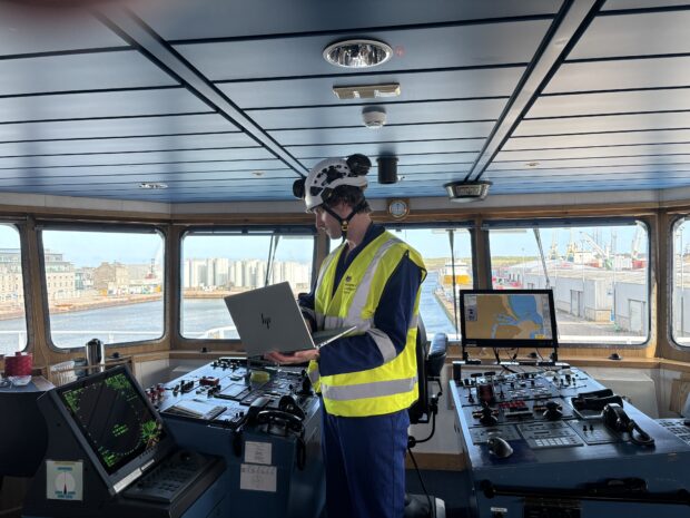 A surveyor in a helmet and high-visibility vest on a ship's bridge holding a laptop at the navigation console.